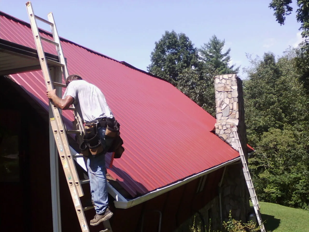 Roofer on a ladder beside a red metal roof and stone chimney — Soffit & Fascia Repair by Roofing Brothers in Haverstraw